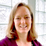 A picture of Susan Jakes, a woman with shoulder-length light brown hair, standing in front of a window and smiling.