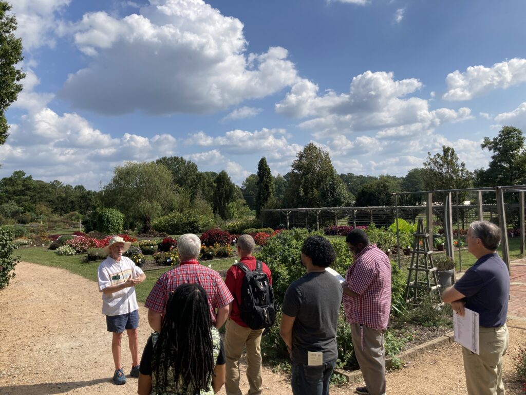 An older man tour guide stands on a gravel path in front of a half dozen Extension employees. They are surrounded by the arboretum gardens on a sunny day.